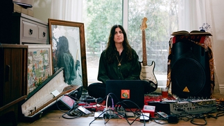Maral sitting in front of a glass door, her fender stratocaster at her back and her studio equipment laid out on the floor in front of her