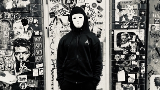 Black and white photo of COEN wearing a white full-face mask and black hoodie in front of a wall covered in posters and stickers