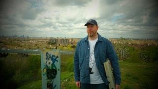 Mr Beatnick standing in a field on a cloudy day. He's wearing an open blue collared shirt, white t shirt and baseball cap, and is holding his MPC sampler under his arm
