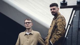 Photo of Blawan and Pariah leaning against an industrial railing while wearing beige jackets