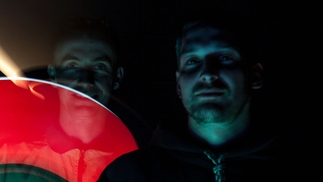 Photo of NKC and DJ Polo standing side by side in a dark room under a grey-blue light. A red segment of light is across NKC's face