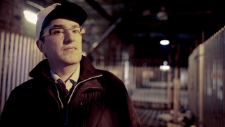 Photo of Marcus Intalex aka Trevino standing in an industrial warehouse. He's wearing glasses, a baseball cap and a black rain coat