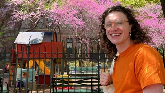 Photo of the producer Compliments smiling in an orange t-shirt. They have long curly brown hair and are wearing glasses and have a tote bag over their shoulder. They're standing in front of a playground and there are cherry blossom trees in the background