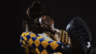 Photo of Little Simz sitting on a chair while wearing a yellow and blue checkered suit