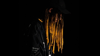 Press shot of Neo Edo on a black backdrop. He's wearing a black baseball cap and is looking down to the right. His long, braided, dyed golden hair covers most of his face