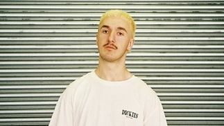 Photo of Franck standing in front of a green corrugated background with bleached blonde hair