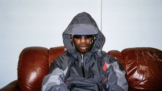 Photo of GAIKA wearing a grey parka and sunglasses while sitting on a sofa
