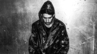 Black and white photo of Catriel Nievas sitting against a wall under a spotlight. His hood is up and he's staring at the ground
