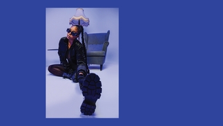 Melanin Tee wearing a black leather coat and boots sitting on the floor infront of a chair. Her hair is braided, she wears sunglasses, and one leg is stretched toward the camera