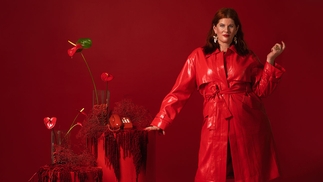 CC:Disco standing in a long red leather coat. She's stood in an entirely red room, next to a red podium with a red telephone on it and red lilys