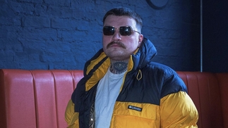 Photo of DJ Deep Heat wearing a yellow puffer jacket and sunglasses in front of a blue wall
