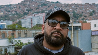 DJ Babatr posing in sunglasses, a black hoodie and black cap, a hilly section of city of Caracas is behind him