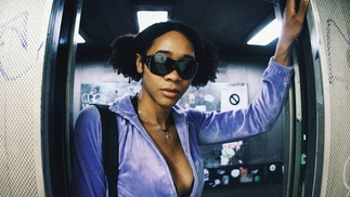Introspekt has her dark hair in space buns, looking straight at the camera from the doorway of an elevator. She rests her hands on either side, and wears bejewelled sunglasses, a purple velour zip up hoodie, a shoulder bag and and blue jeans. 