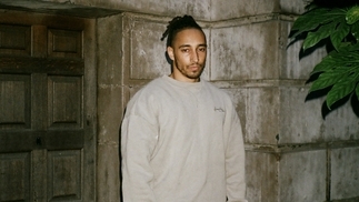 Otik wearing a grey hoodie in front of a stone building with plants