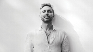 Black and white photo of David Guetta posing against a white wall in a white open collar shirt