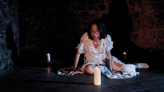 gyrofield sits on a wooden floor in a cave-like room next to a candle wearing a distressed white dress