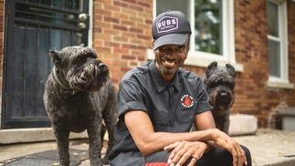 Amp Fiddler sitting outside of his home in Detroit, smiling next to his two dogs