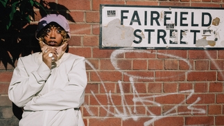 Nia Archives poses in an anorak in front of a red brick wall with white graffiti on and a sign that reads "Fairfield Street"