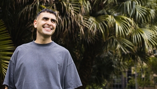 Sammy Virji standing in a washed blue t-shirt in front of plants. He is smiling and looking away from the camera.