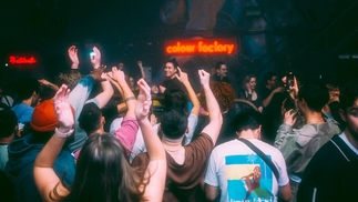Photo of Danielle DJing taken from the dancefloor in Colour Factory. The club's sign hangs behind her and people in the crowd have their hands in the air