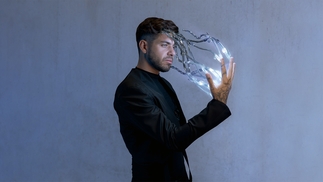 Anyma press shot, showing standing in front of a blue-grey wall in a suit, with electricity travelling between his fingers and his face