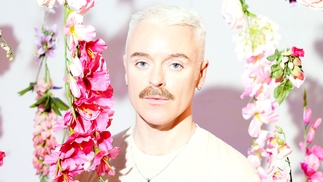 A brightly lit photo of Cormac in a white t-shirt next to a variety of pink hanging flowers