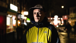 Photo of Earl Grey wearing a yellow and navy raincoat and baseball cap. He's standing in a city street at night