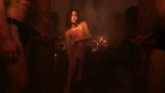 Elkka wearing a pink dress in a steamy room filled with candles. A person in tight black briefs is toward the front of the photo on the left