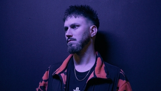 Photo of Mani Festo wearing a black and red collared fleece, standing against a dark wall under a purple hued light