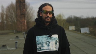 Assyouti standing outside near a train track on an overcast day, wearing sunglasses and a Slint hoodie