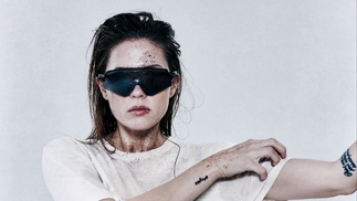 Charlotte De Witte wearing a white t-shirt and shades, her left arm is held aloft to her left, while she scratches her left shoulder with her right hand