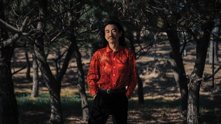 DJ Nobu wearing a red button up shirt and black trousers, standing outside amongst some trees