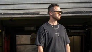 Photo of Pattn wearing a black t-shirt and sunglasses in front of an industrial background
