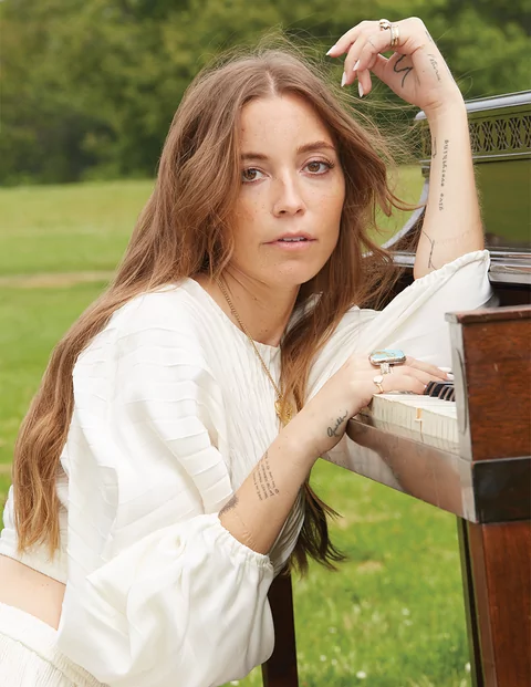 Photo of LP Giobbi wearing a white long-sleeved dress and resting her arms on a piano in a field