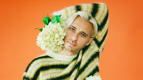 A press shot of Flume in a striped jumper, holding a bunch of white flowers against an orange backdrop 