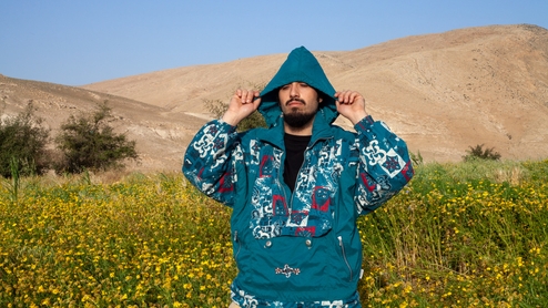 Julmud stands in a field of yellow flowers, in front of a mountain and blue sky, holding the hood of his blue sweater over his eyes