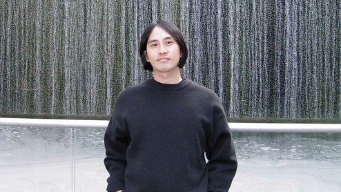 Stones Taro standing in a black sweater in front of a wall
