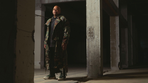 DJ Bone in a long army camo jacket standing in a concrete doorway