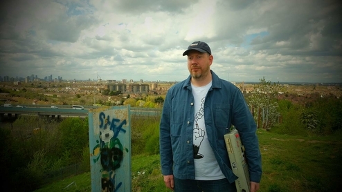 Mr Beatnick standing in a field on a cloudy day. He's wearing an open blue collared shirt, white t shirt and baseball cap, and is holding his MPC sampler under his arm