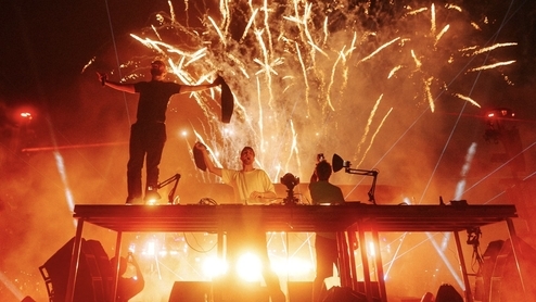 Photo of Skrillex, Four Tet, and Fred Again performing at Coachella with orange fireworks. 