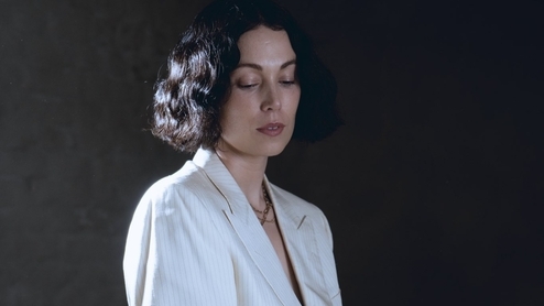 Photo of Kelly Lee Owens sitting behind a mossy foreground wearing a white suit