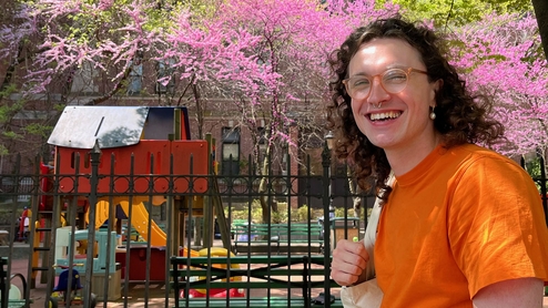 Photo of the producer Compliments smiling in an orange t-shirt. They have long curly brown hair and are wearing glasses and have a tote bag over their shoulder. They're standing in front of a playground and there are cherry blossom trees in the background