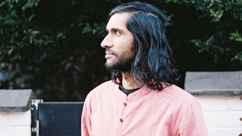 Balraj Singh Samrai looking to the left of the camera. He's in front of a low wall with a leafy tree above it. He's wearing a pinkish red collarless shirt. his black hair is shoulder length 