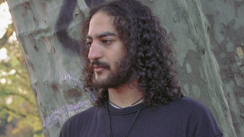 Photo of Assyouti standing in front of graffiti-covered pillar. His hair is long and curly and has a beard.