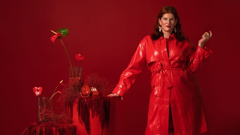 CC:Disco standing in a long red leather coat. She's stood in an entirely red room, next to a red podium with a red telephone on it and red lilys