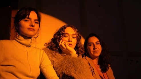 LOTTIE PENDLEBURY, LENNY WATSON, SOPHIE FARRELL of SISTER MIDNIGHT sat against a wall with evening sun shining on them through a window