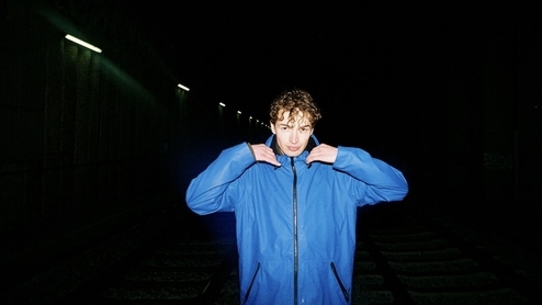 Photo of DJ JM wearing a blue parka jacket with a dark-lit industrial background