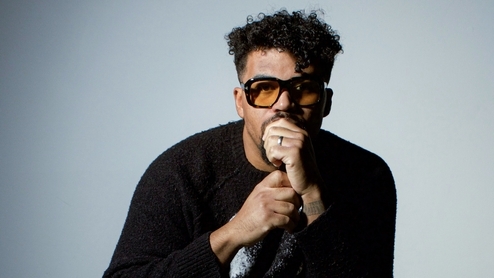 Photo of DJ Cuddles wearing orange sunglasses and a black sweatshirt in front of a light blue background