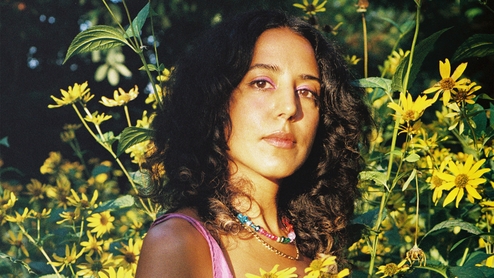 Photo of Ayesha wearing pink eyeshadow and colourful jewellery in front of a large bush of yellow flowers