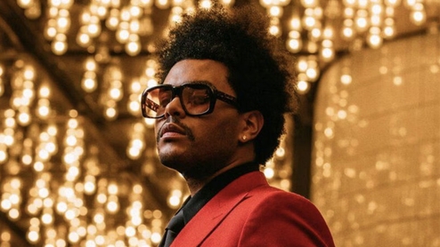The weeknd poses in a room filled with hanging lights. He's wearing a red suit and large sunglasses